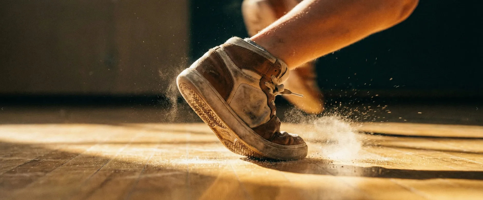 A ground-level, extreme close-up of a child's foot in a sage green sneaker and oatmeal sock, frozen mid-sprint on a wooden floor. Dust particles are kicked up at the point of impact, visually representing the 'Speed' pillar of First Pick's scientific 5+3 development methodology