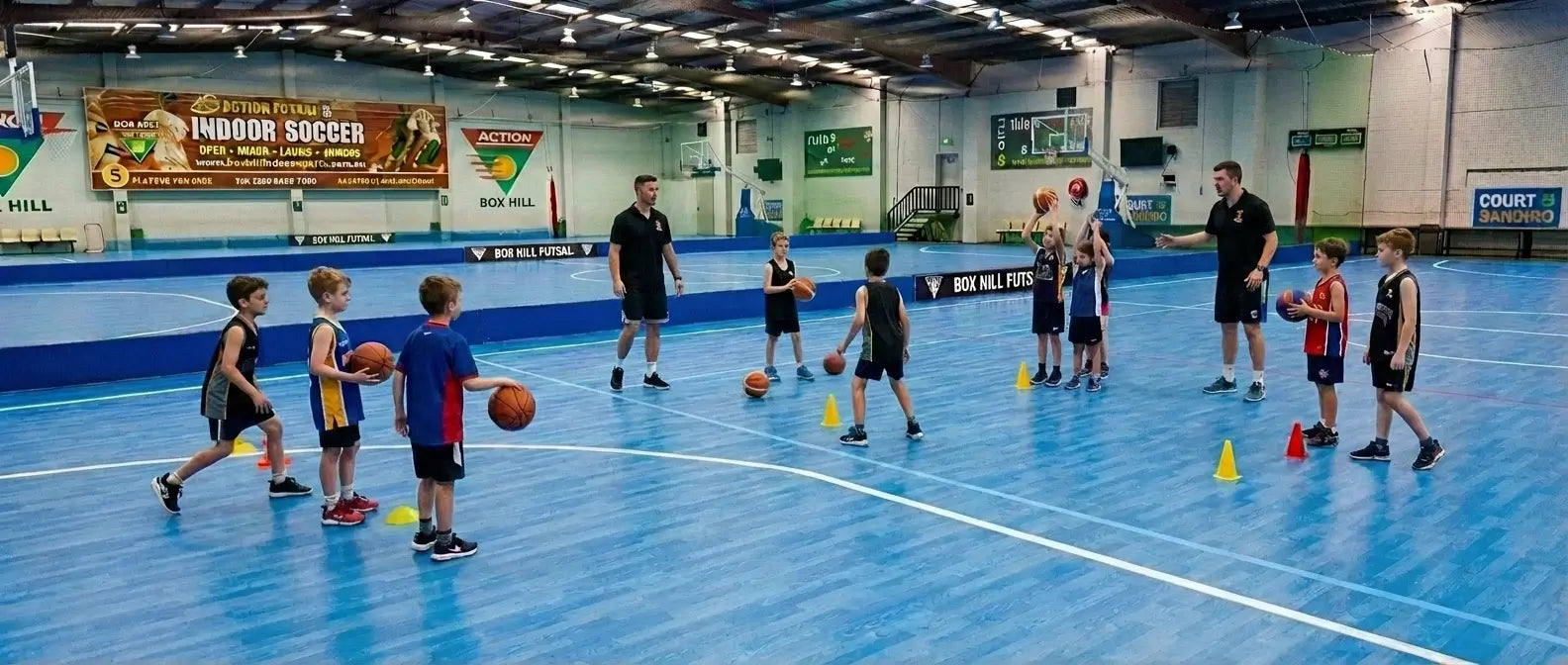 A high-energy junior basketball training session at First Pick Academy's Box Hill South campus, showing elite coaches working with children and 5 years old basketball beginners on a professional blue court.