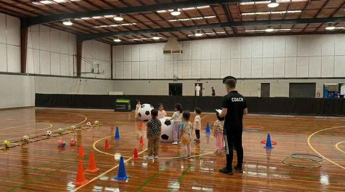 A professional basketball coach in a black uniform working with young children at First Pick Academy's indoor Melbourne facility, using specialized sports equipment to build early childhood focus and movement skills.